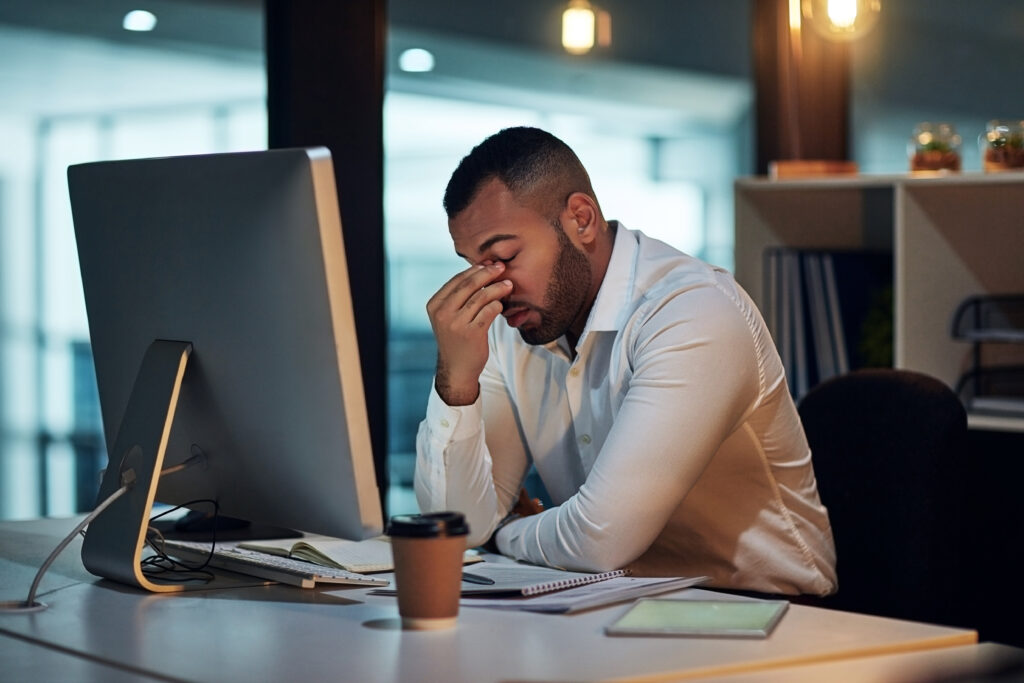 man pinching his nose in frustration while sitting in front of a desktop monitor at an office desk.