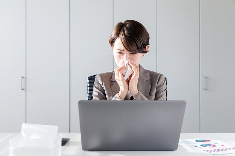 A woman uses a tissue in front of a laptop. Workplace Productivity––How Your HVAC Plays a Role.